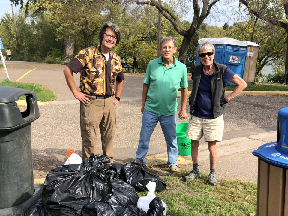 Three smiling people standing outdoors next to a parking lot at Como Lake in Saint Paul. There is a large pile of full black trash bags in front of them on the grass, in between waste and recycling bins. 