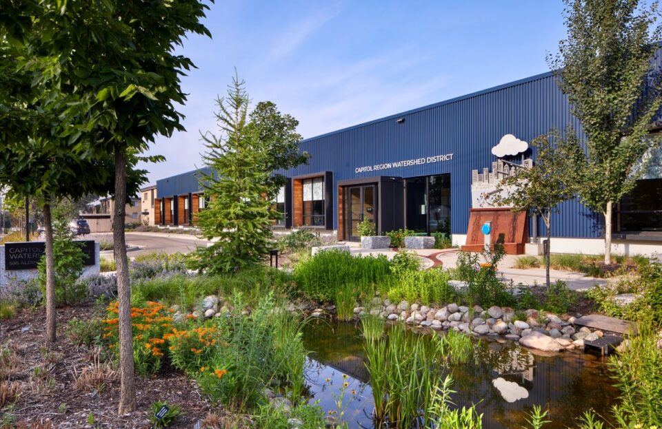 A pocket park with a small pond surrounded by native plants. An interactive exhibit with a rain cloud and city skyline in front of a modern, one-story office building is in the background.