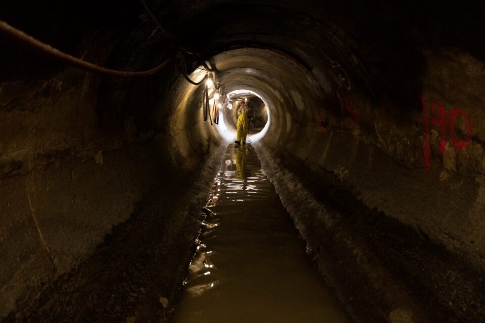 A worker wearing a hard hat, headlamp, and waterproof overalls walks in a few inches of water in the bottom an 8-foot diameter concrete storm sewer tunnel. The number 40 is spray painted in bright pink on the right wall of the tunnel and lighting is strung along the opposite side. 