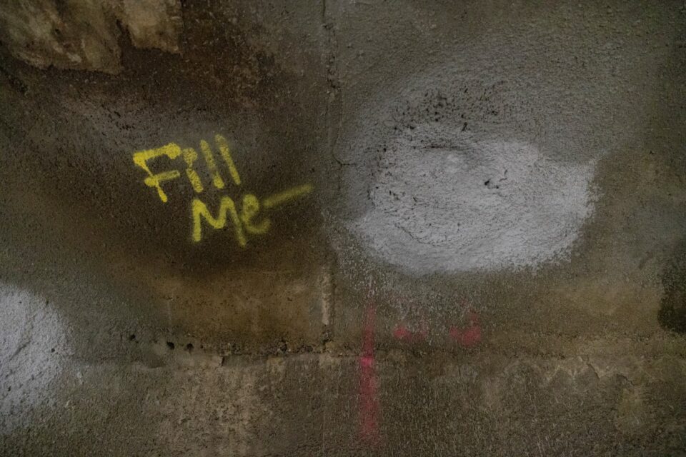 A freshly repaired hole covered in spray concrete on a patched concrete wall. Next to the hole it says ‘Fill Me’ in yellow spray paint. 