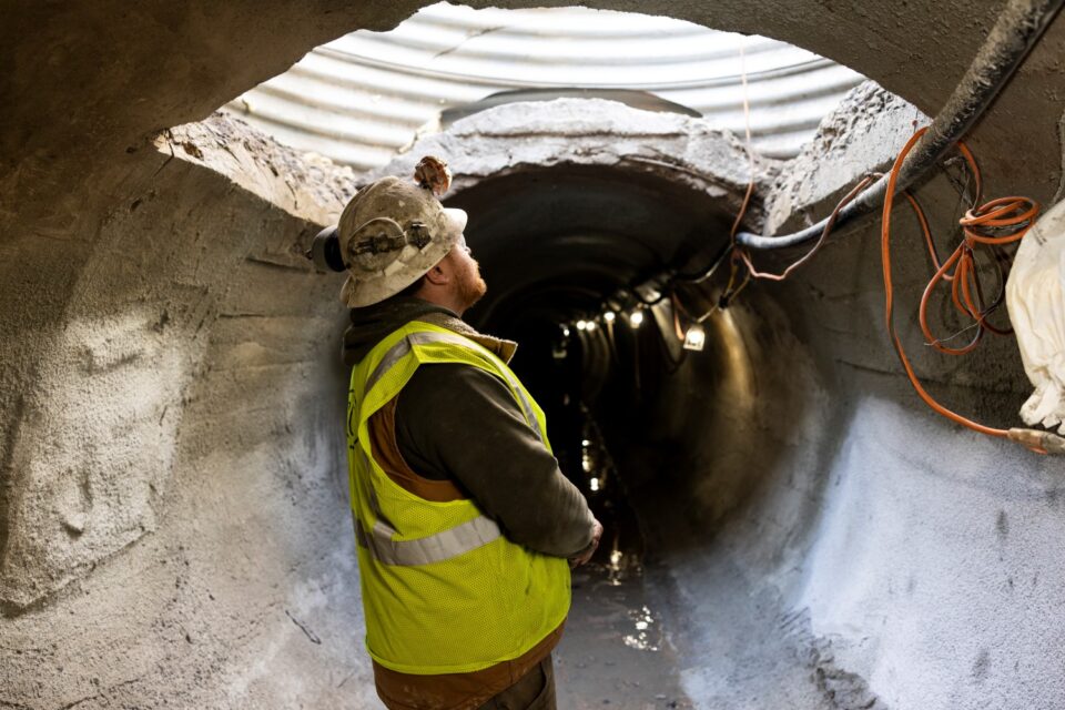 A person in a hard hat and safety vest looks up in an 8-ft diameter storm sewer tunnel with a large opening cut into the top. 