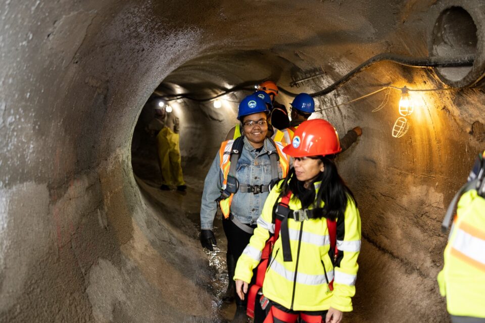 Women wearing hard hats, eye protection and safety vests and harnesses walk single file in an 8-ft diameter concrete storm sewer tunnel. The tunnel is lit by a string of light bulbs along one side. 