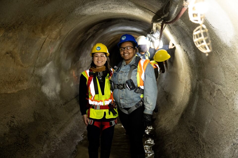 Saint Paul City Councilmembers Hwa Jeong Kim and Cheniqua Johnson stand in an 8-foot diameter concrete storm sewer. Both women are wearing a hard hat, eye protection, and a safety vest and harness. 