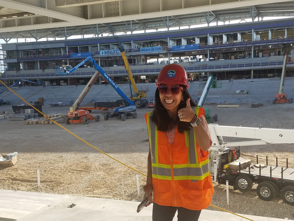Anna Eleria is wearing a safety vest and a hard hat while standing, giving a thumbs up sign, and smiling in front of a large stadium construction site with various machinery and construction materials visible.