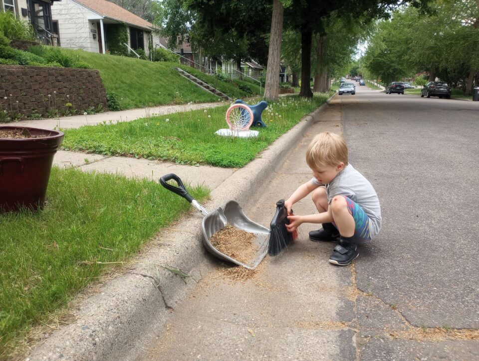 A child crouched on the ground sweeping brown leaves and pine needles from a gutter into a dustpan on a residential street. There is a yard bin and a tipped-over children’s basketball hoop on the boulevard, with parked cars down the street in the background.