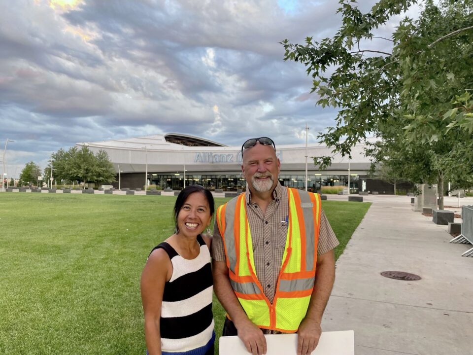 Anna Eleria and former Administrator Mark Doneux are smiling and standing in front of Allianz Field stadium. Anna wears a black and white striped top and Mark wears a fluorescent safety vest over a brown short-sleeved button-up shirt. Behind them is a cloud-streaked sky and well-maintained grass lawn and greenery around a paved walkway.