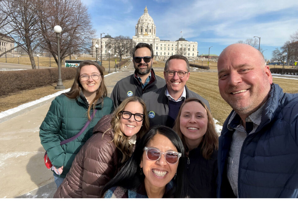 A group of seven smiling adults taking a selfie on a sunny day with the Minnesota State Capitol building in the background. They are wearing winter coats and jackets. The lawn and the walkway behind them are well-maintained, with some snow on the dormant, brownish grass and bare trees along the path. 