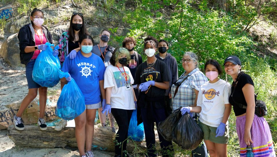 A group of eleven volunteers, some wearing masks and gloves holding full, blue and black trash bags. Behind them are large rocks and bright green trees and bushes. 