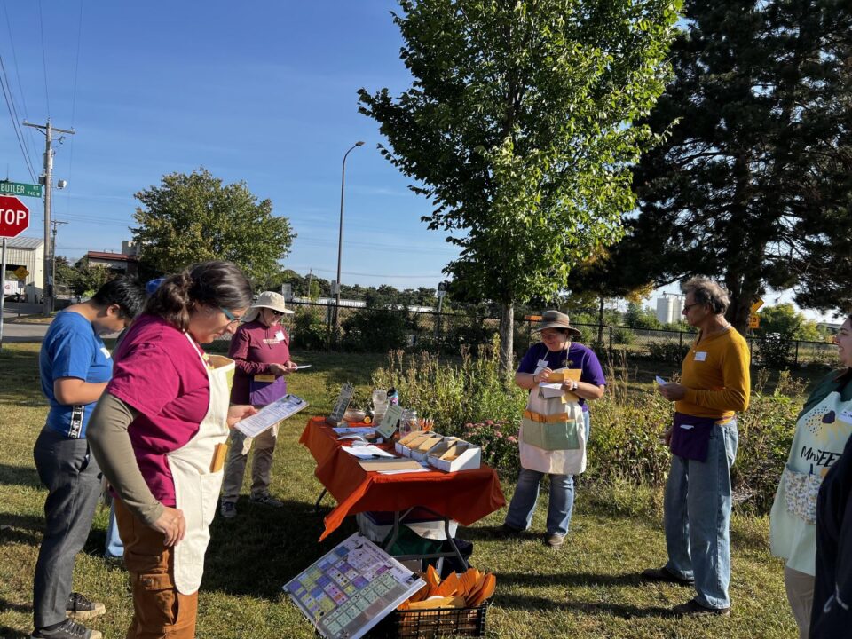 Outdoor community gathering with several people standing around a table covered with informational materials. Two individuals are holding papers, and another is engaged in conversation. A 