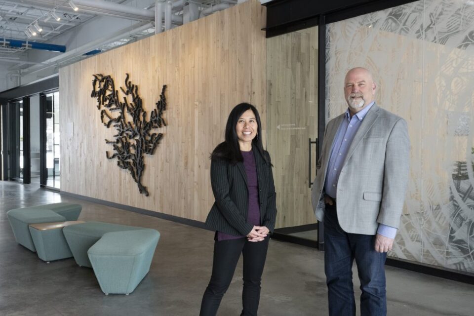 Anna Eleria and former Administrator Mark Doneux are smiling and standing in Capitol Region Watershed District's lobby. Behind them, a wood-paneled wall has an intricate iron art piece showing the Mississippi River watershed.