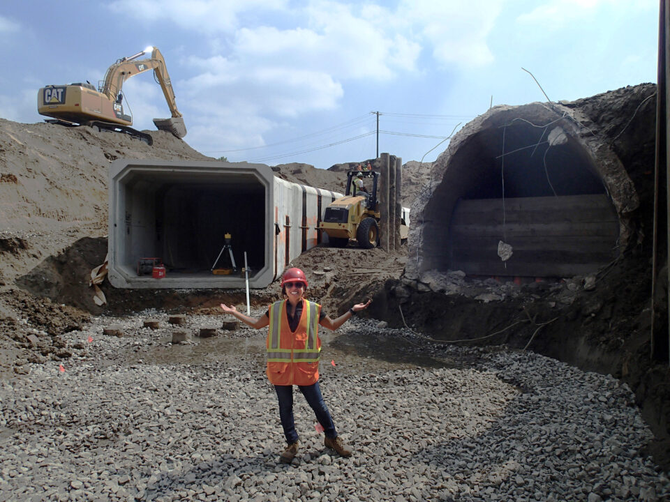 Anna Eleria smiles in a safety vest and hard hat, standing with arms extended in a large construction site with two tunnel sections, a forklift and a backhoe in the background. The tunnel section on the right is horseshoe-shaped and has a concrete wall in its entrance to block it. The tunnel on the left is brand new and rectangular-shaped. 