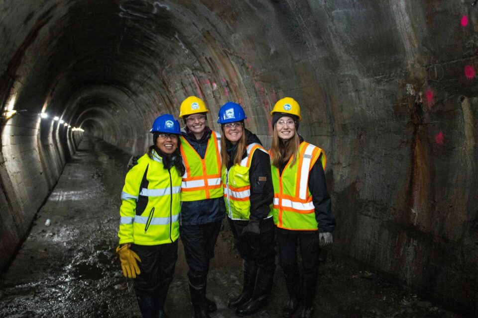 Anna Eleria and three other women, all wearing hard hats with a CRWD logo and high-visibility vests, smile while standing together in a large, arched storm sewer tunnel. The concrete tunnel walls with pink survey markings are visible behind them.