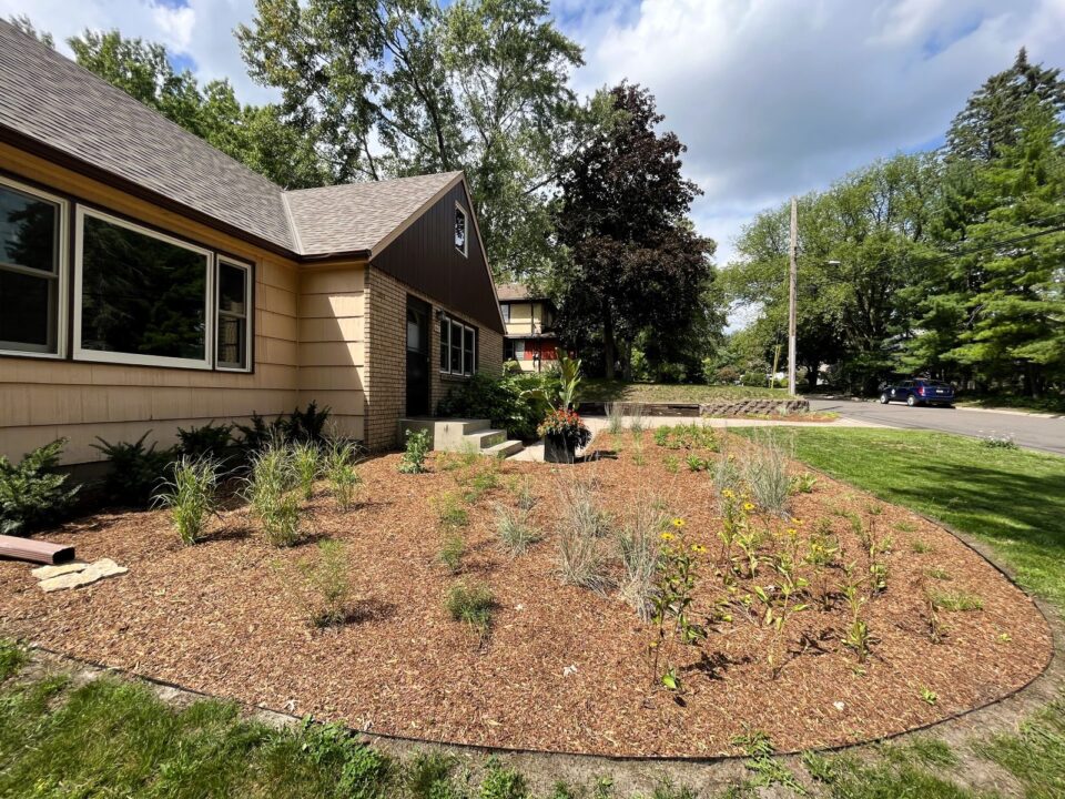 Landscaped garden in front of a single-story house with a variety of native plants in a freshly mulched circular bed. 