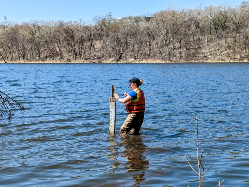 A person wearing waders and a life vest standing in shin-deep water in Crosby Lake next to a level gauge with lines and numbers to measure the lake’s water level on a post. 