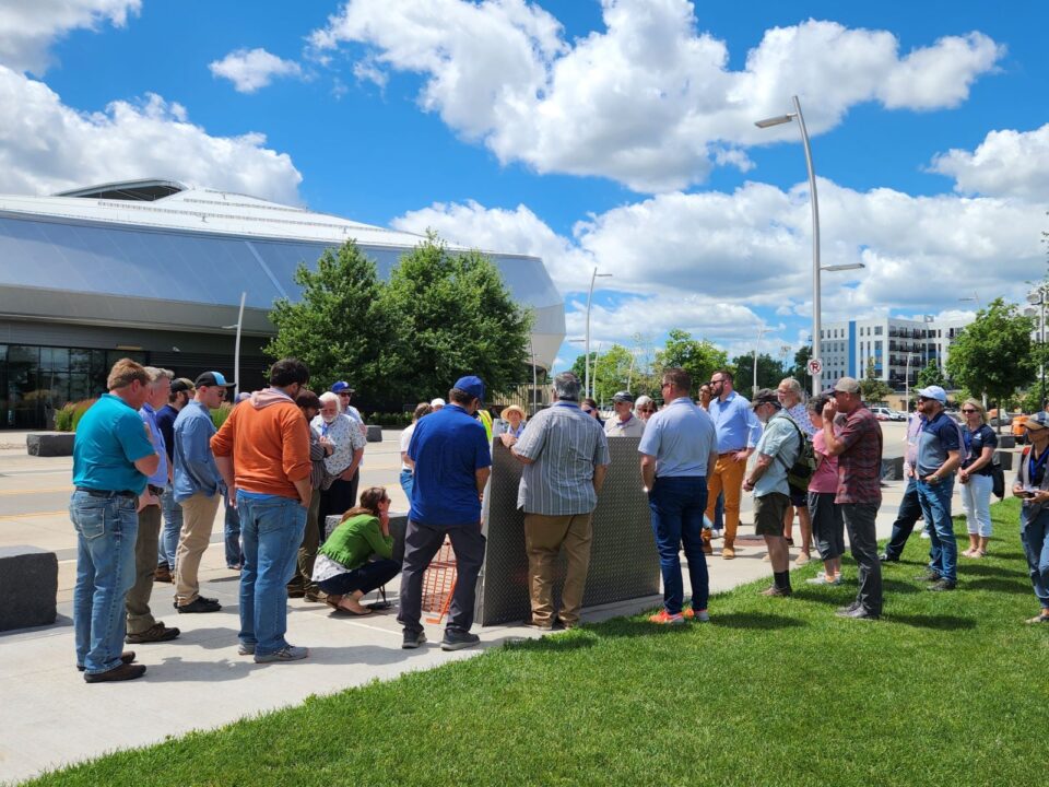 A group of people are standing, and one is crouched around an open hatch on the sidewalk in front of the modern, angular Allianz Field under a bright blue sky with puffy clouds. 