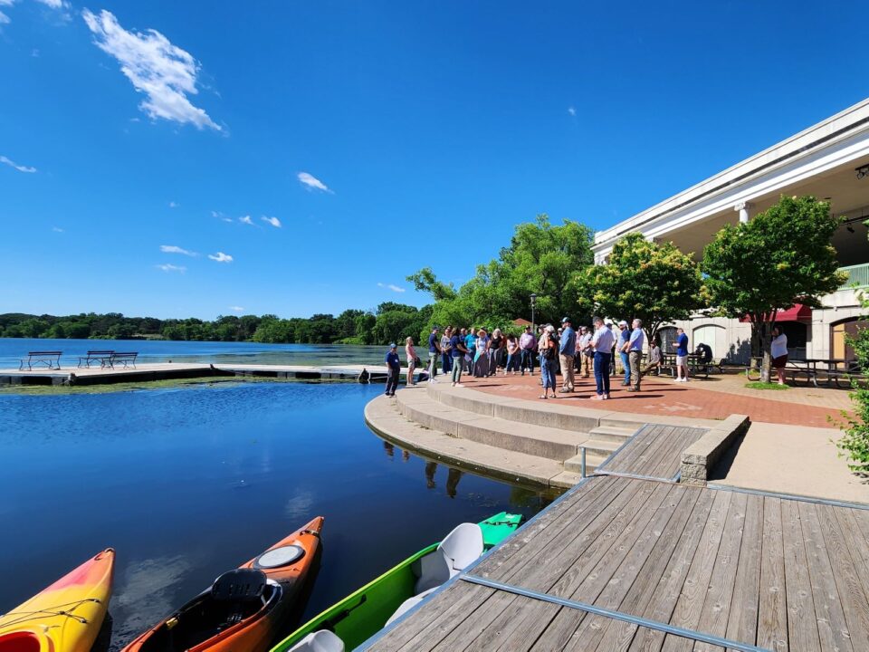 On a sunny day, a group of people outside the Como Lakeside Pavilion, with colorful kayaks on the dock by the lake reflecting the blue sky.