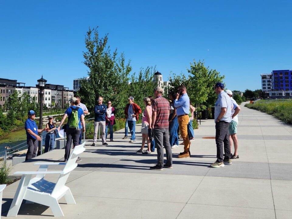 A group listening to a tour guide on a paved path along the Highland Bridge water feature with modern buildings and green landscaping in the background. 