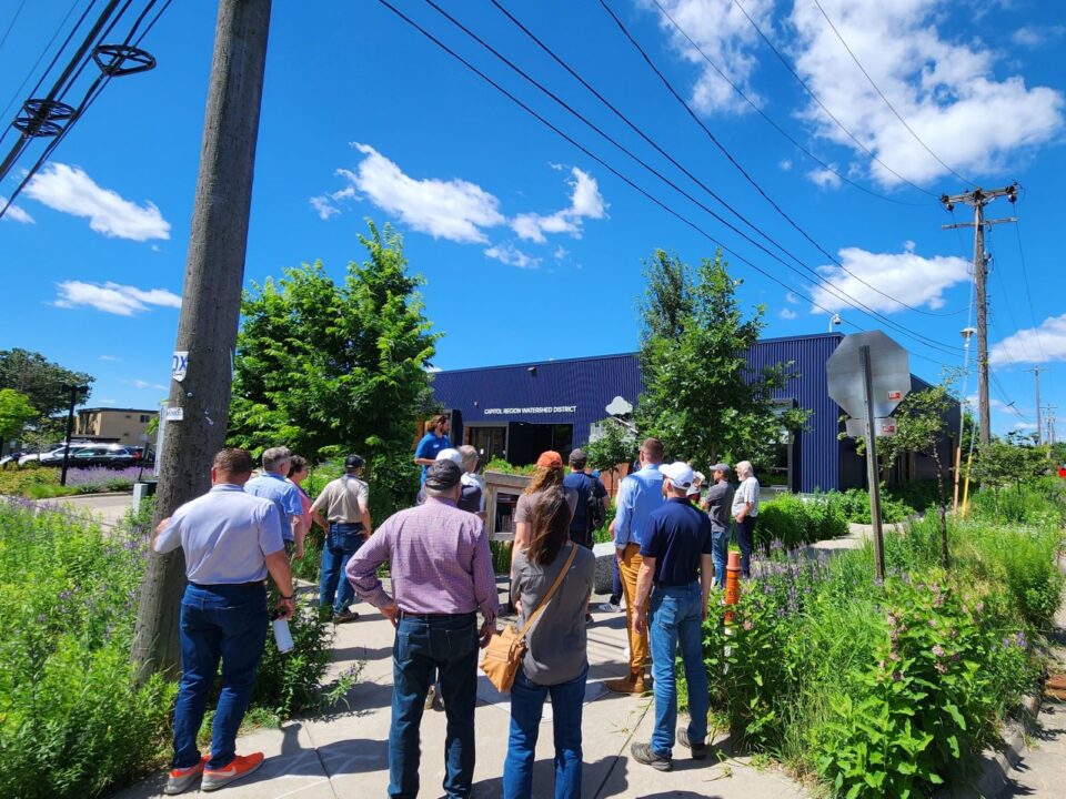 A group is in front of CRWD's one-story office building at a corner of the pocket park under a clear blue sky, listening to a tour guide. Native plants and trees surround the park's sidewalks. 