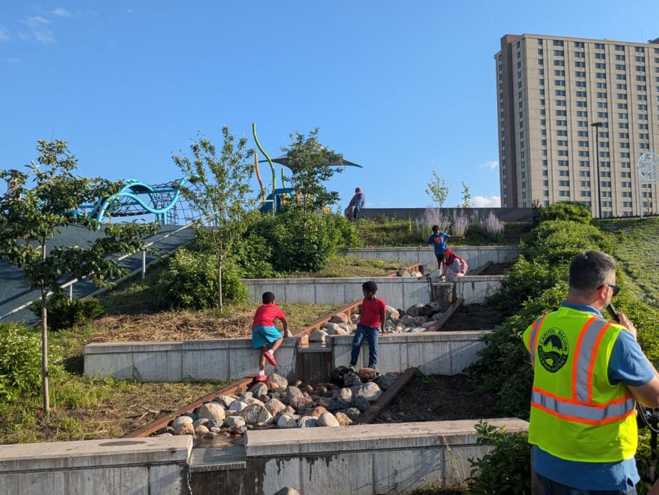 Children playing on a rock-lined, terraced water feature surrounded by greenery as an adult looks on. The setting is in a park with a playground in the background. In the foreground, someone in a safety vest with the Capitol Region Watershed District logo is speaks to someone outside of the frame.  