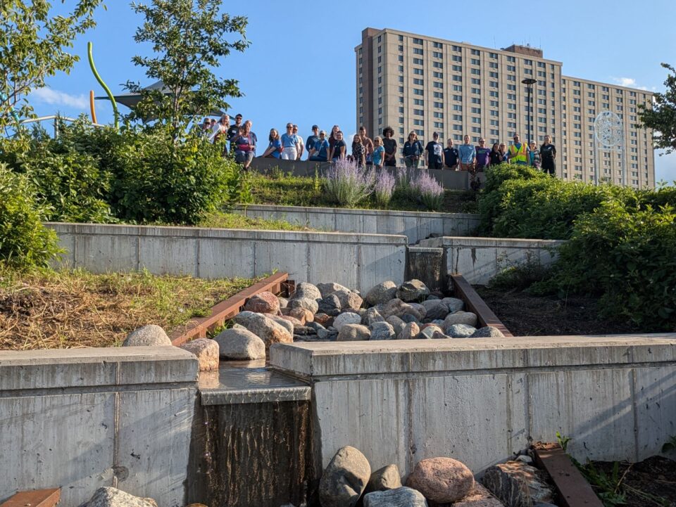 A group of people standing on a landscaped terrace overlooking a rock-lined water feature. In the background, a large multi-story building under a clear sky.