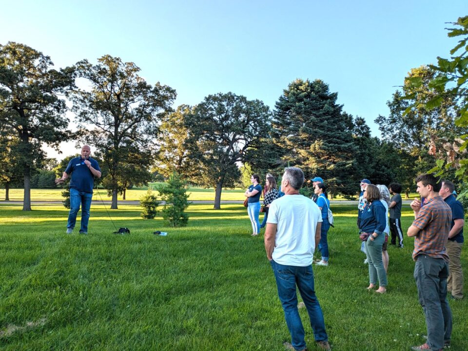 Group of people standing in a park, attentively listening to a speaker who is addressing them on a bright sunny day. The setting includes expansive green lawns and scattered trees.