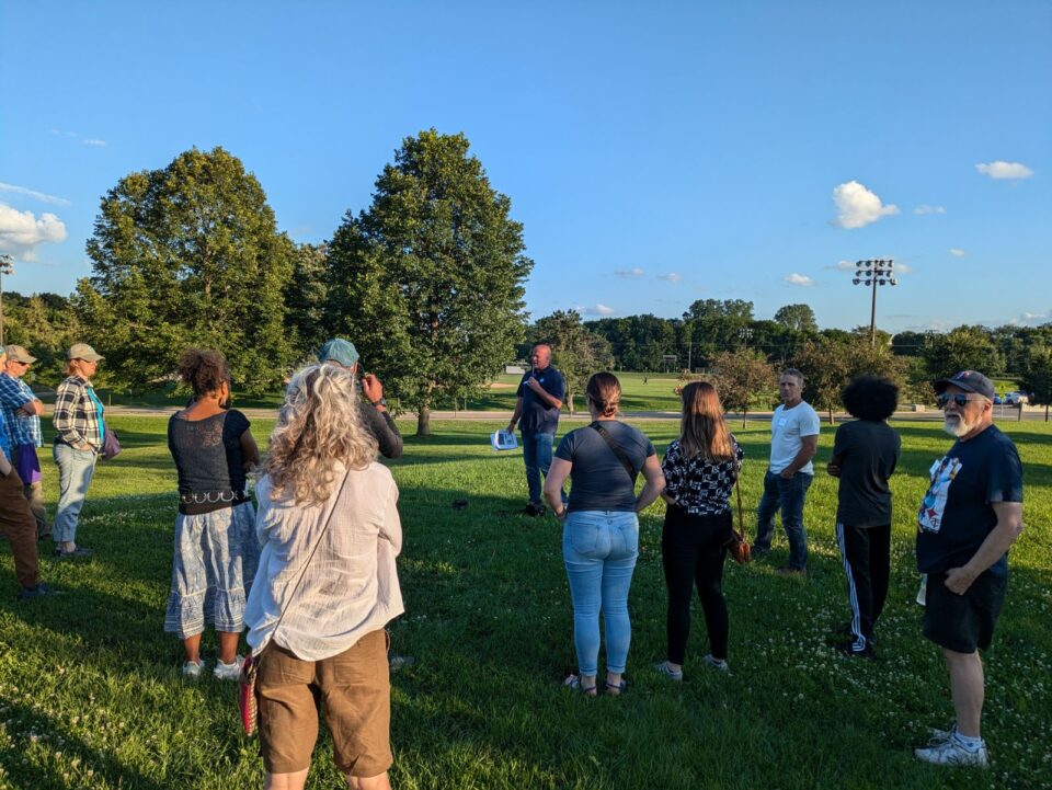 A group of people standing in a park listening to a person giving a presentation. Baseball fields and a large field light post are in the background.