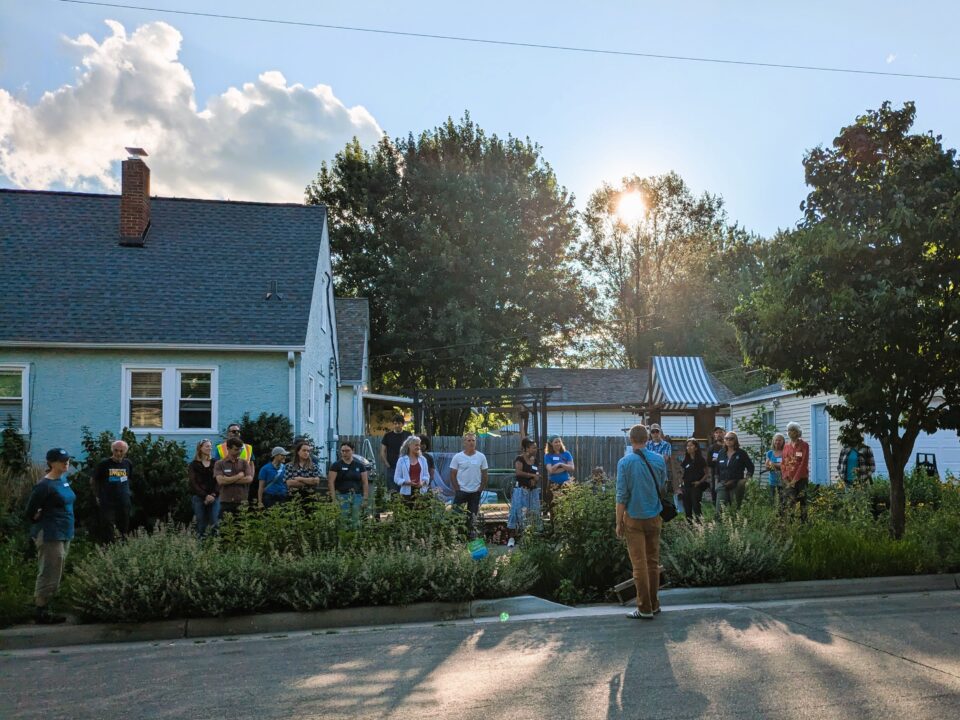 A group of people gathered along a sidewalk next to a house, listening to a speaker standing in the street. The boulevard between them is full of green plants, flowers, and a tree, there is a 2-foot-wide break in the curb leading to the garden. Sunlight filters through the trees, creating a serene ambiance.