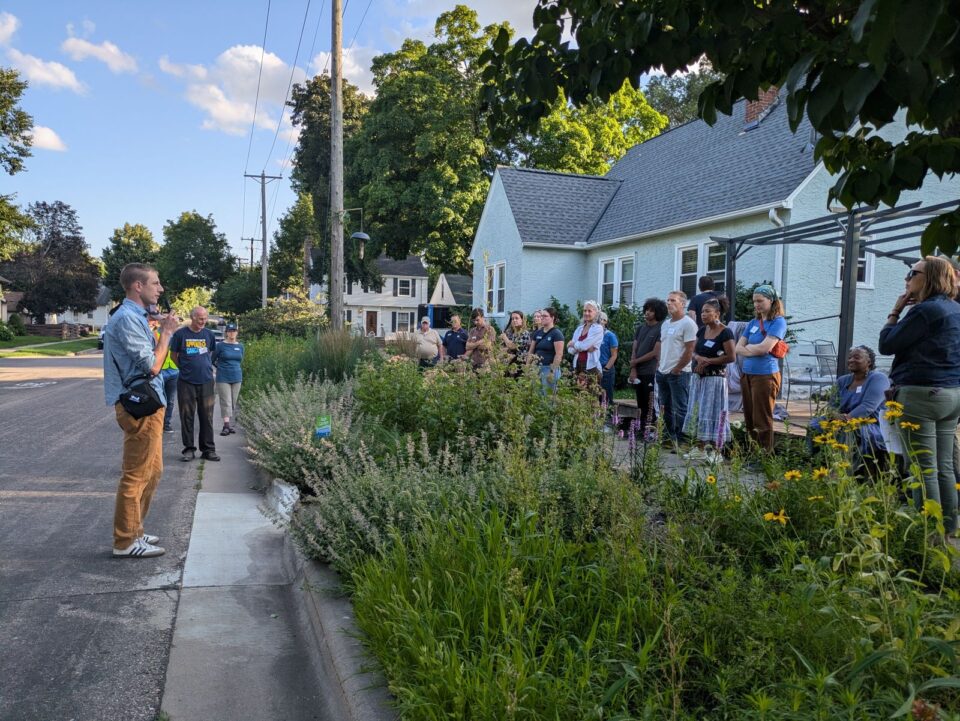 A person stands addressing a group of attentive listeners gathered on a residential street with lush greenery of native plants under a clear sky.