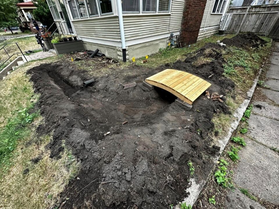 Freshly dug trenched garden along the front corner of a residential home’s yard. There is a small wooden foot bridge over the garden and a buried downspout ending in the garden’s edge. 