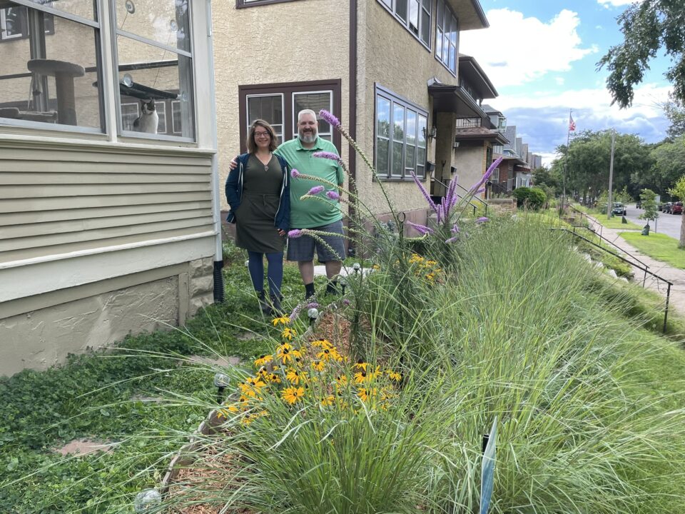 Jen and Mike standing in between their house and a rain garden full of tall grasses and blooming native plants with long, spiky purple blossoms on tall arched stems and yellow petaled flowers with a dark brown center. 