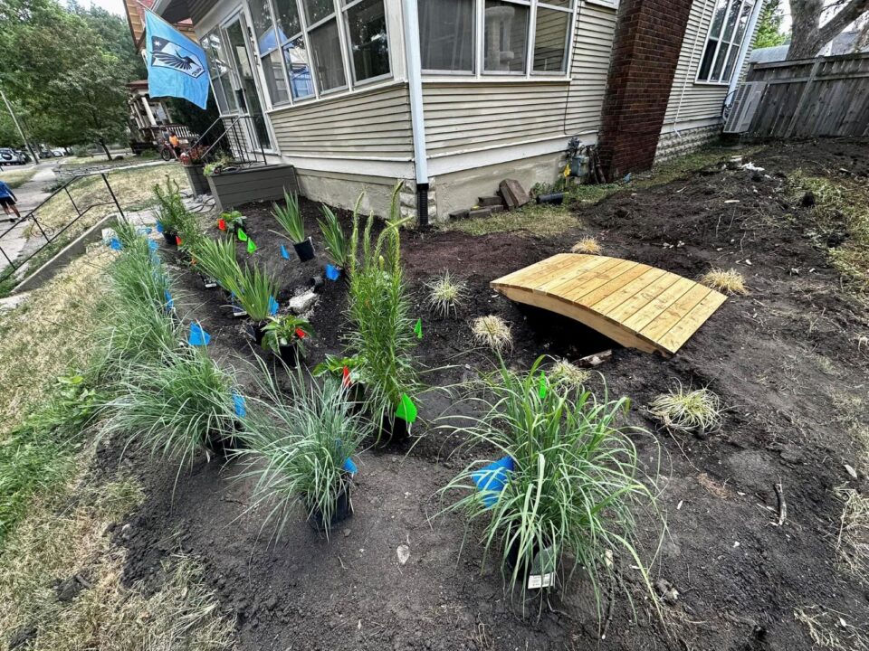 Front yard of a house with a freshly dug garden, and a row of native grasses and plants in starter pots placed next to blue, green and red landscaping flags. 