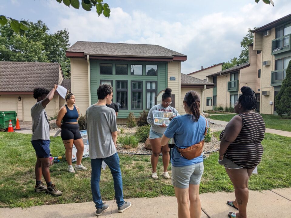 A group of people standing in a loose circle outside the Hampden Square townhome office, engaged in a conversation while one person holds a sign with a grid of native plant photos.