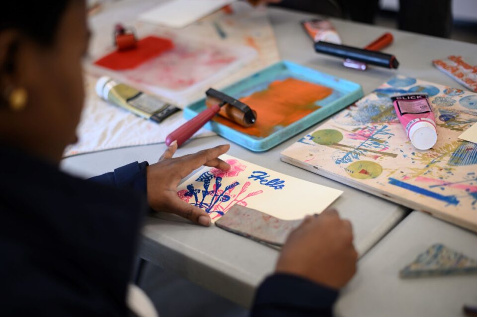 Person creating a colorful print using a stamps at a table scattered with art supplies.