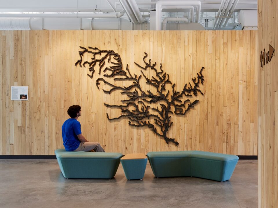 A person sitting on a blue bench, looking at a large iron sculpture of the Mississippi River watershed on a wood paneled wall in a modern office.  