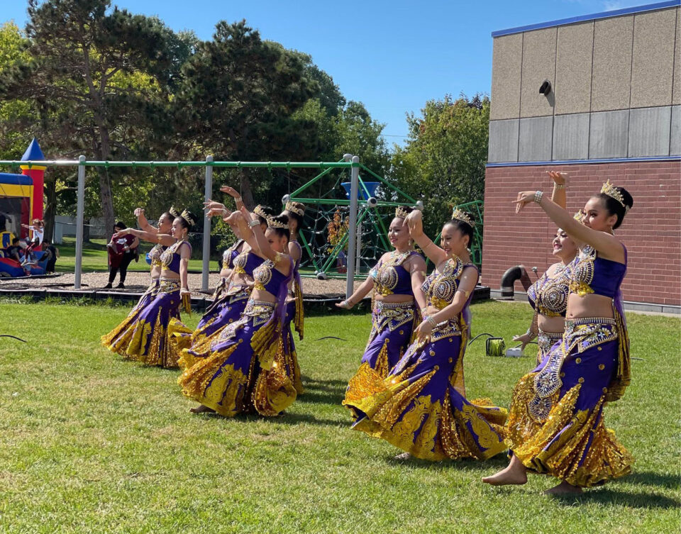 A group of dancers perform in colorful traditional costumes of purple and gold with lots of embellishments at an outdoor event on a field of green grass beside a playground.