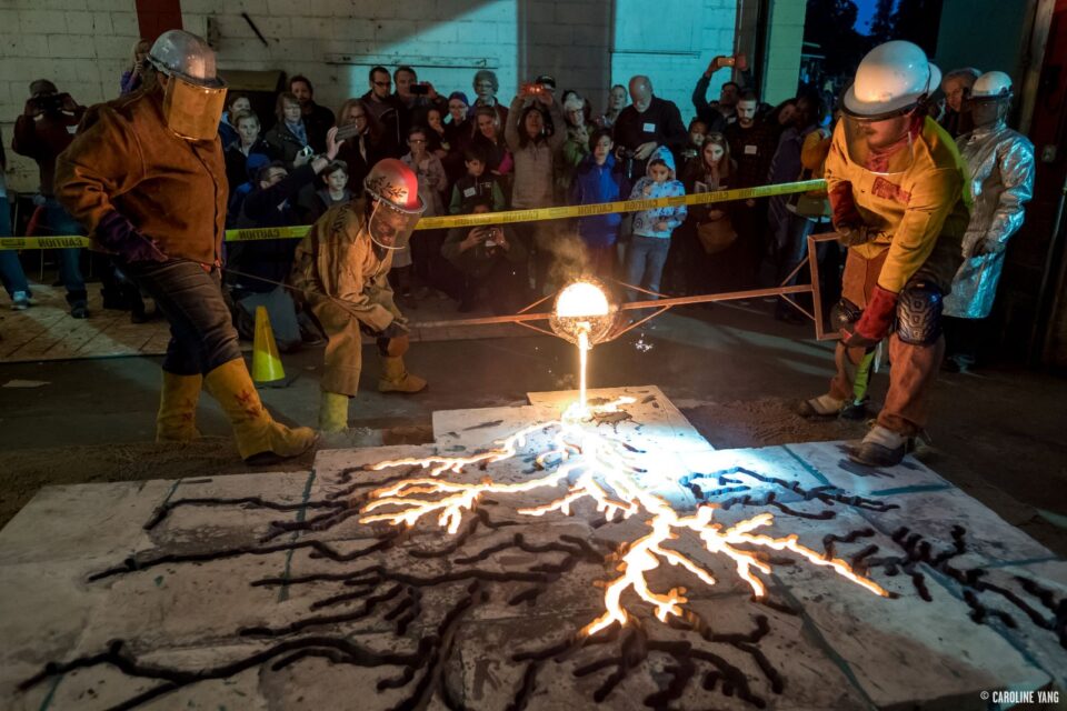 Three people in heat safety gear, helmets and face shields stand around a large cast of the Mississippi River watershed laying on the ground. Two people pour glowing, molten iron into the cast by holding each end of a long pole with a metal bucket suspended in the center that contains the iron. A crowd of people watches them from behind a line of yellow caution tape. 