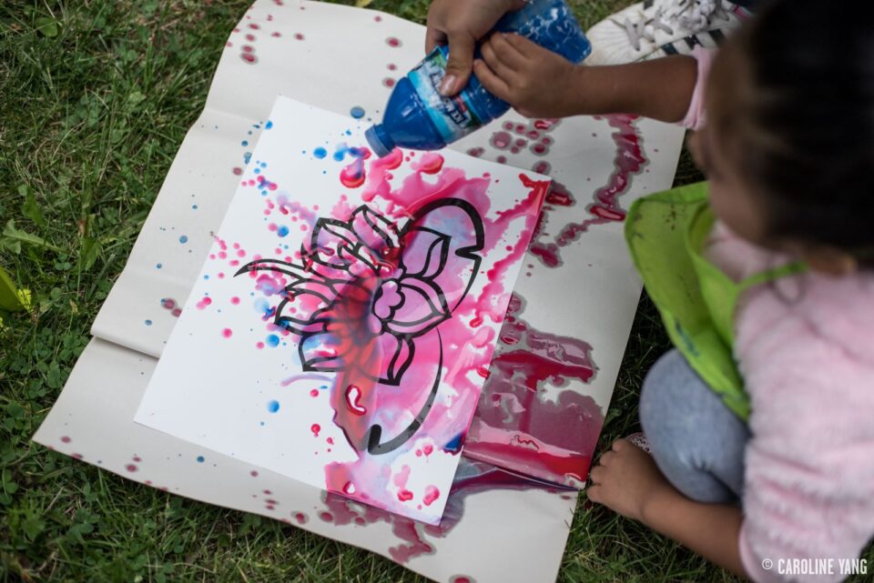 A child squeezing colorful paint from a plastic water bottle on to a drawing of a water lily, while another child watches.