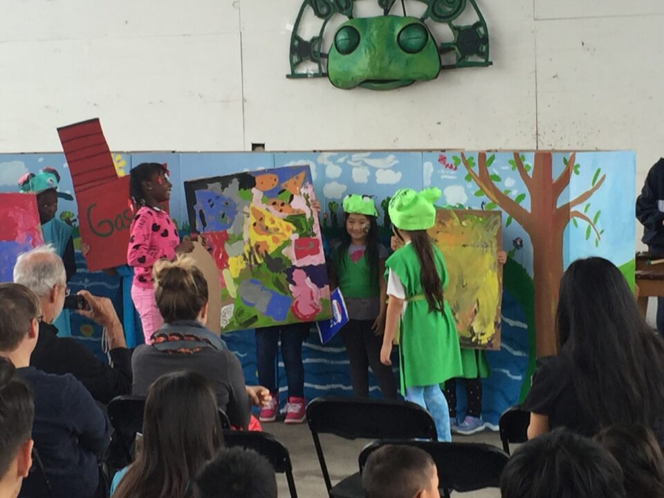 A group of children perform for people sitting on folding chairs. The children are wearing bright clothes and some wearing paper-mache frog masks in front of a hand painted backdrop of a lake with blue sky and trees. There is a large paper-mache frog face on the wall behind them. 