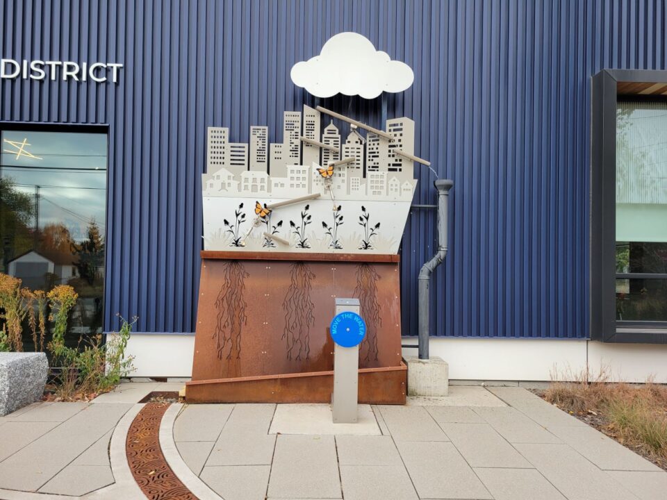 A sculptural metal exhibit outside of a modern office building. A city skyline with a cloud cutout above, orange butterflies, cutouts of native flowers above rusted metal with root cutouts. A metal post with a bright blue wheel is in front of the exhibit.