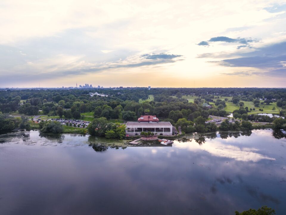 Aerial view of the west side of Como Lake with the Como Lakeside Pavillion, a white building with a red roof. The calm lake reflects the sky with scattered clouds at sunset. The distant downtown Minneapolis skyline is on the horizon. 