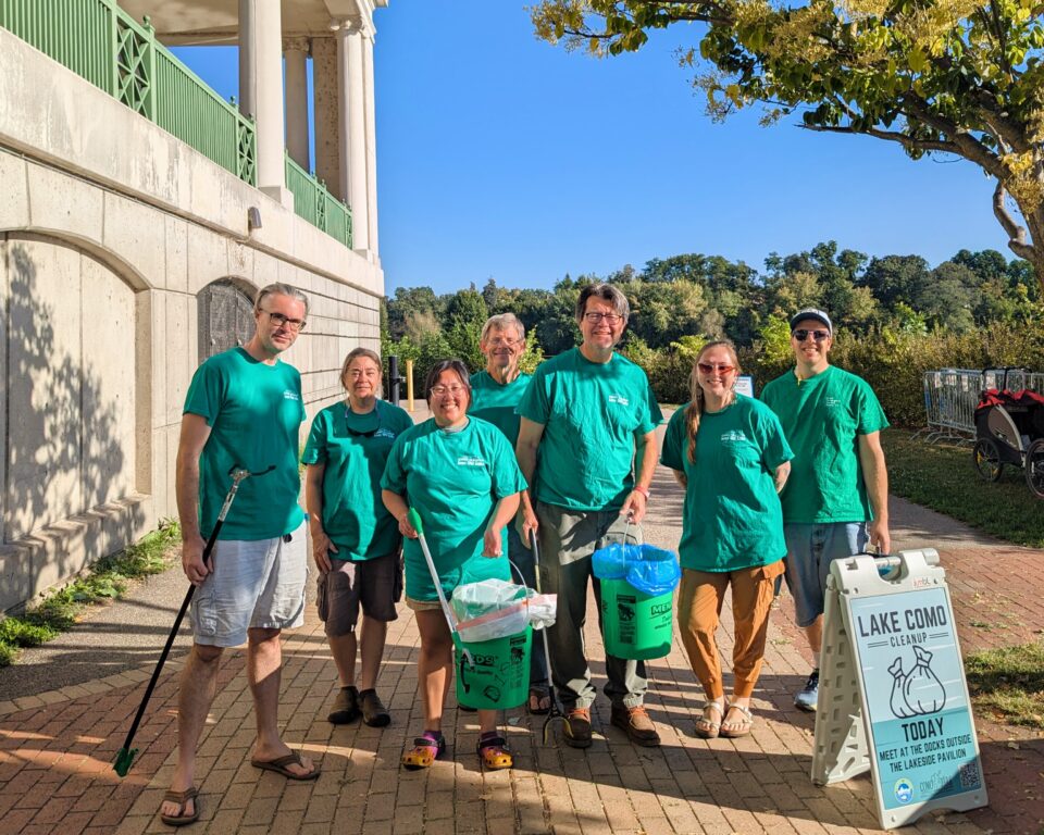 Seven smiling people in matching green shirts standing with trash grabbers and five gallon buckets near a sign that reads 