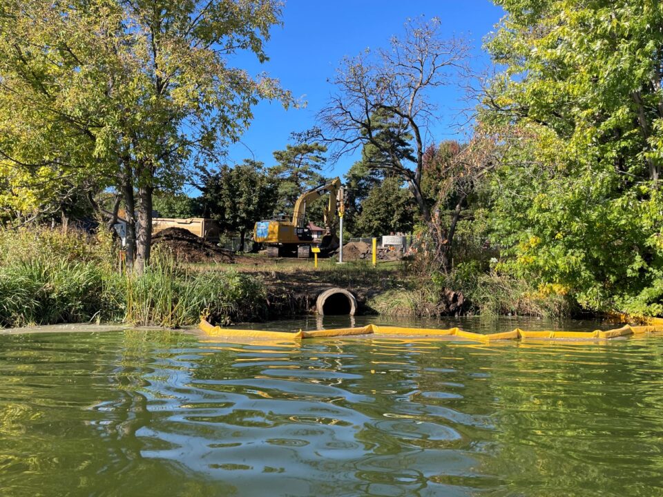 An excavator and construction equipment above a storm sewer outfall, a concrete pipe in the lakeshore, at Como Lake. A yellow containment boom in front of the outfall floats in the lake. 