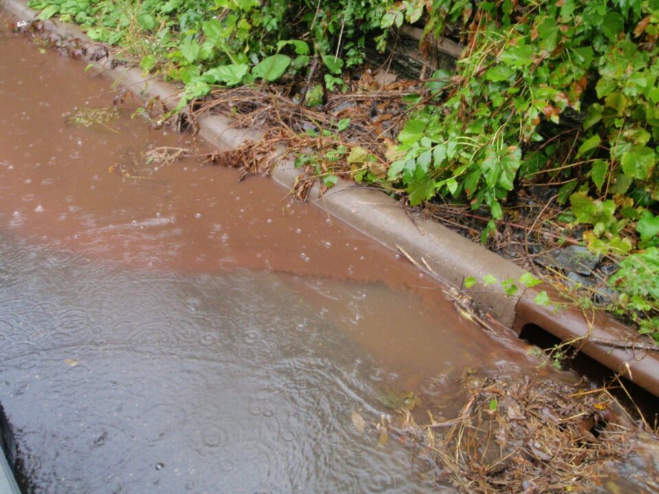 Muddy water with erosion from a construction site flows along a curb to a storm drain during a rainstorm. The drain is covered with leaves and debris.