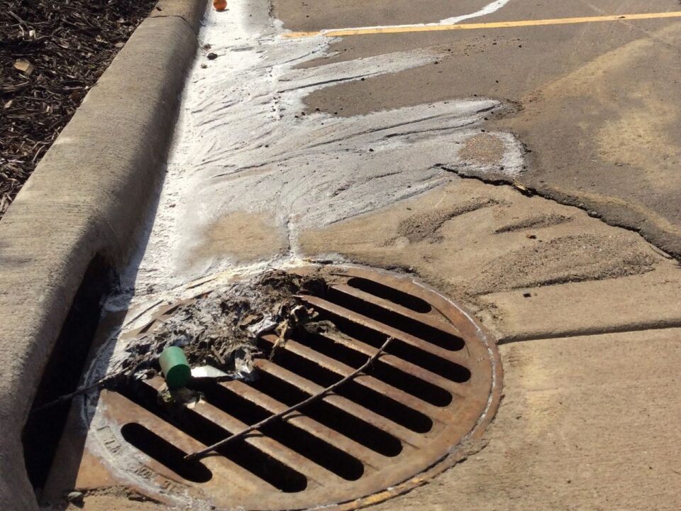 A parking lot storm drain with debris on the grate, and a patch of dried, white spill leading to the drain from across the parking lot.. 