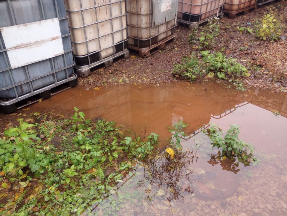 Large industrial containers sit on a muddy ground with pools of murky water and patches of green vegetation.