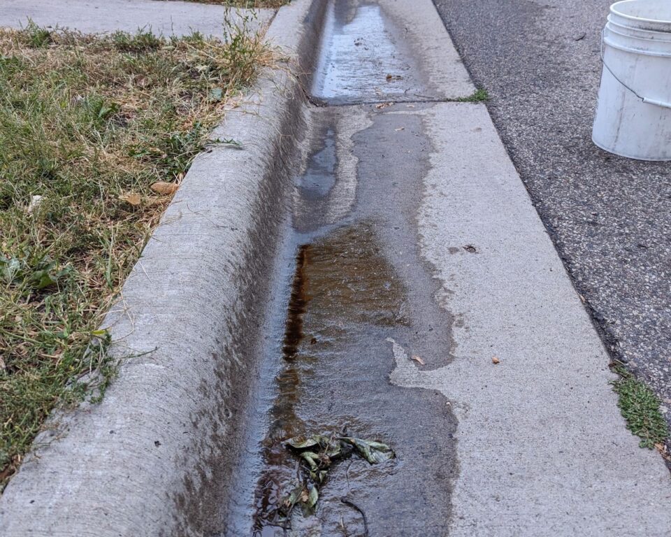 Dark brown motor oil pools in water flowing along a street curb.