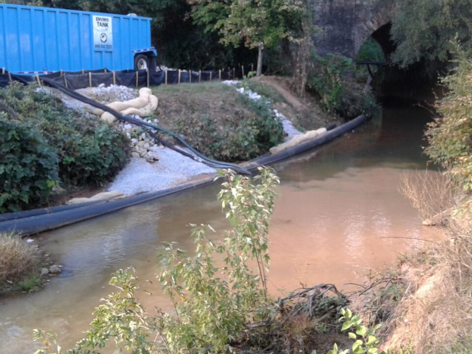 A stream with muddy water draining from a large hose running down the stream bank from a blue truck in a construction site. Trees and vegetation are on both sides of the stream with an old stone tunnel in the background. 