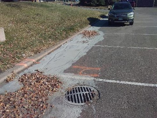 A drainage grate in a parking lot with a large plume of chalky, gray clay from water that flowed to the drain. A pile of brown leaves is on the side of the drain, also covered in gray clay. A car is parked in the background.  