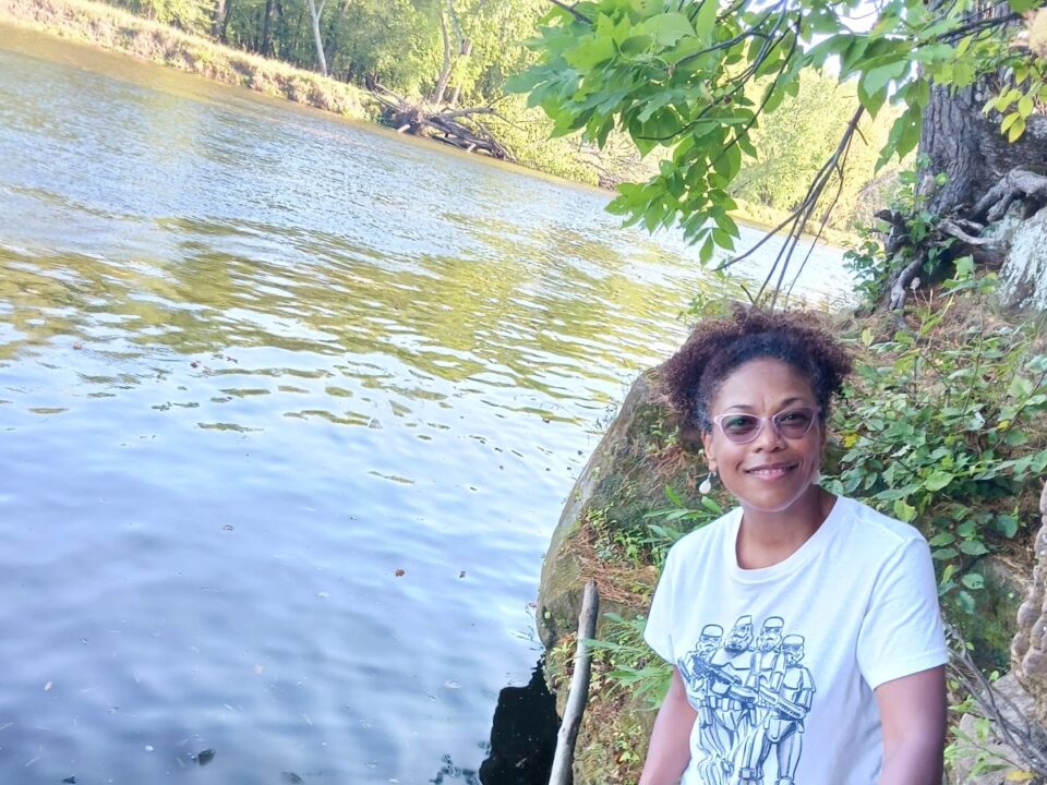 A woman standing along a serene riverbank. She is smiling and wearing sunglasses and a T-shirt. 