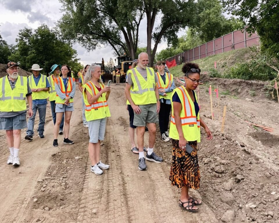 Group of people in dayglo yellow and orange safety vests touring a construction site with piles of dirt and excavation equipment in the background.